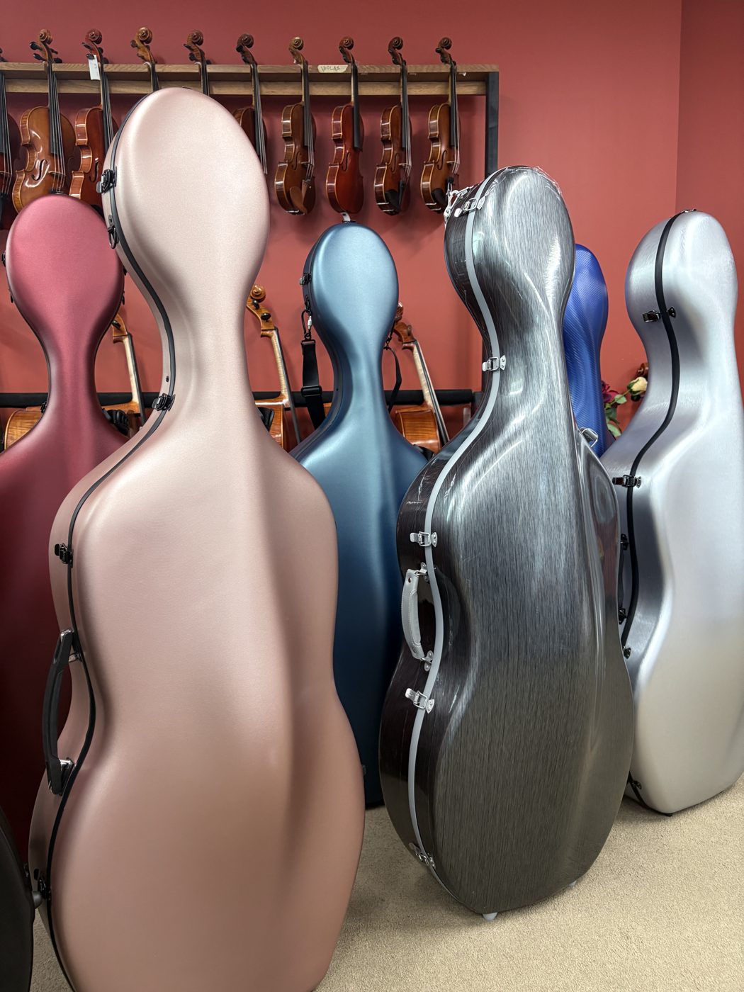 Cello cases in pink, grey, blue and silver standing in front of violas hanging on the wall.