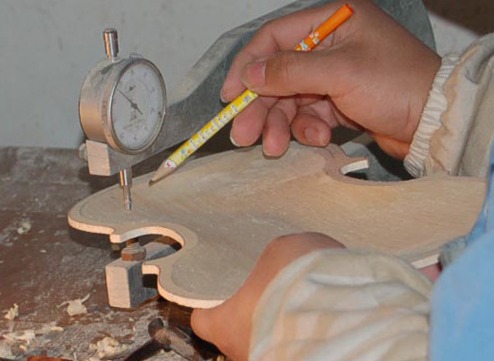 A maker marks a violin plate in pencil while checking its thickness with a dial gauge.